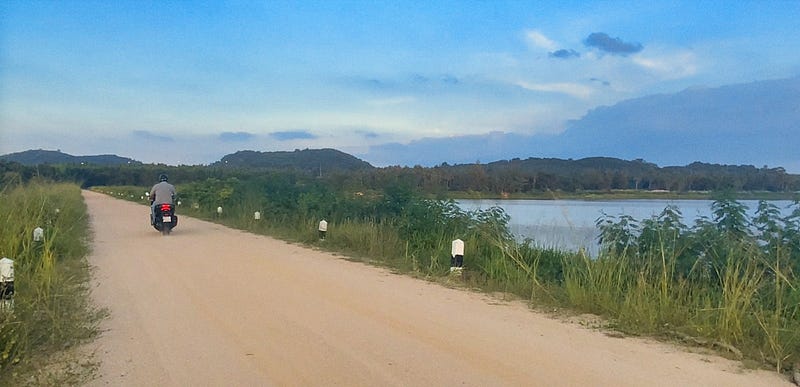 A black Honda Forza parked on a quiet gravel road beside a lake, southern Thailand, late afternoon light